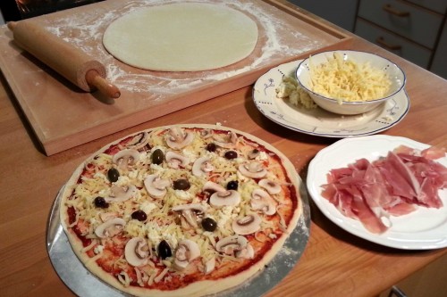 Brödet i botten, sedan tomatsåsen och osten, sist påläggen. Fin skinka läggs på när pizzan har bakats nästan klar.